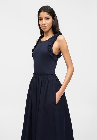 Γυναίκα που φοράει φόρεμα σε σκούρο μπλε χρώμα χωρίς μανίκια με πλισέ τιράντες, εφαρμοστή κορσέ, σούρα στη μέση και φουσκωτή φούστα με τσέπες, στέκεται μπροστά σε απλό φόντο.