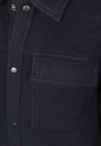 Donker marineblauwe wollen jas close-up met zichtbare drukknoopjes, een klepzak en gedetailleerde stiksels op een textuurstof.