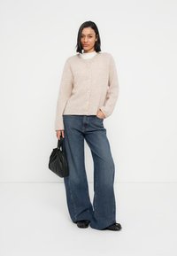 Ανοιχτό μπεζ ζακέτα με κουμπιά από απαλό πλεκτό υλικό, σε συνδυασμό με πλατύτερο τζιν παντελόνι σε μπλε χρώμα. Το μοντέλο κρατάει μια μαύρη τσάντα.