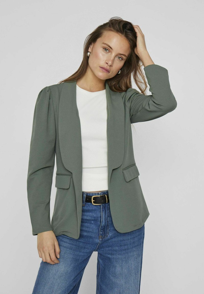 Blazer verde con una silueta estructurada, que presenta solapas anchas, dos bolsillos frontales y una tela texturizada, combinado con una blusa blanca y jeans azules.