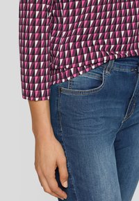 Hand ruht neben blauen Denim-Jeans und trägt ein langärmeliges Oberteil mit geometrischem Muster in Purpur, Schwarz und Weiß.