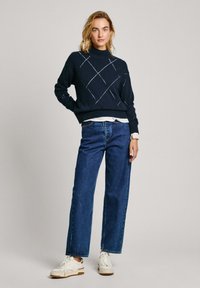 Marineblauwe gebreide trui met ruitpatroon, hoge hals en geribbelde manchetten, gecombineerd met losse blauwe spijkerbroek en witte sneakers.