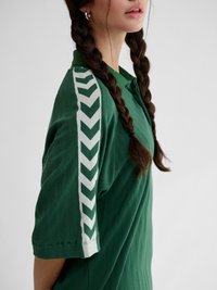 Ung kvinne med to flettede hårstrenger iført en grønn poloshorte med hvite chevron-striper på ermene.