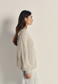 Svetlo sivi pleteni pulover z ohlapnim krojem, rebrasto teksturo in spuščenimi rameni. Kombiniran s svetlim denimom, viden od strani na nevtralnem ozadju.