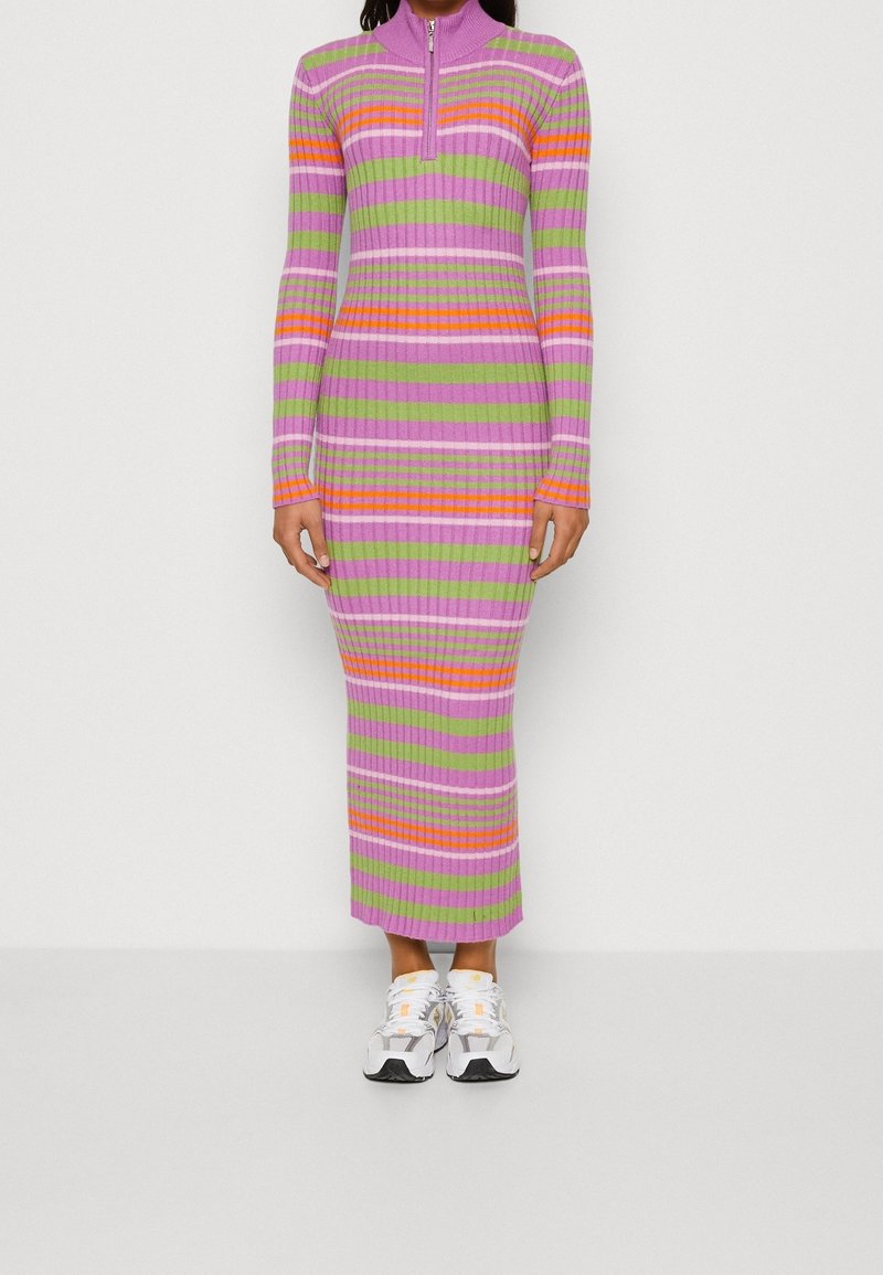 Femme portant une longue robe en maille côtelée avec des rayures horizontales violettes, vertes, orange et rose clair, associée à des baskets blanches.