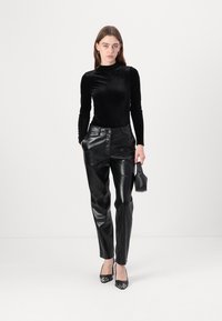 Haut en velours noir à manches longues associé à un pantalon en cuir noir taille haute. Le modèle tient un petit sac et porte des talons noirs.