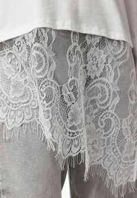Tissu en dentelle florale blanche recouvrant un pantalon gris clair avec un haut blanc, affichant des motifs de dentelle délicats, complexes et des bords frangés.