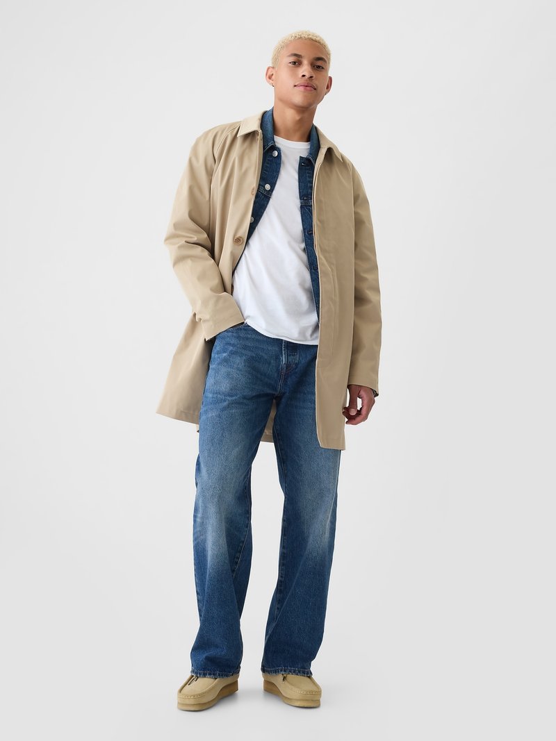 Beige Trenchcoat über einer Denimjacke, kombiniert mit einem weißen T-Shirt, blauen Jeans und beigen Schuhen. Glatter Stoff und entspannte Passform.