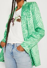 Blazer verde texturizado con un acabado sedoso, que presenta mangas largas y un cuello con muescas. Llevado sobre una camisa blanca y combinado con jeans claros.