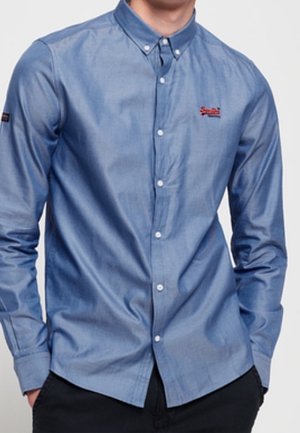 Mężczyzna ubrany w niebieską koszulę z długim rękawem z małym czerwonym logo na piersi, z rękami w kieszeniach czarnych spodni.