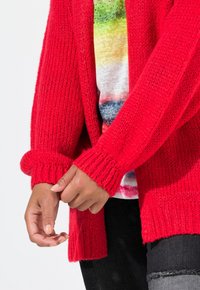 Cardigan rosso lavorato a maglia con maniche lunghe, dotato di polsini a coste e apertura frontale, indossato sopra una maglia multicolore. Dettaglio del tessuto struttura visibile.