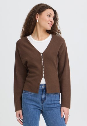 Vrouw met krullend haar die een bruine knoopvest draagt over een wit hemd en blauwe spijkerbroek, kijkt naar de zijkant tegen een effen achtergrond.