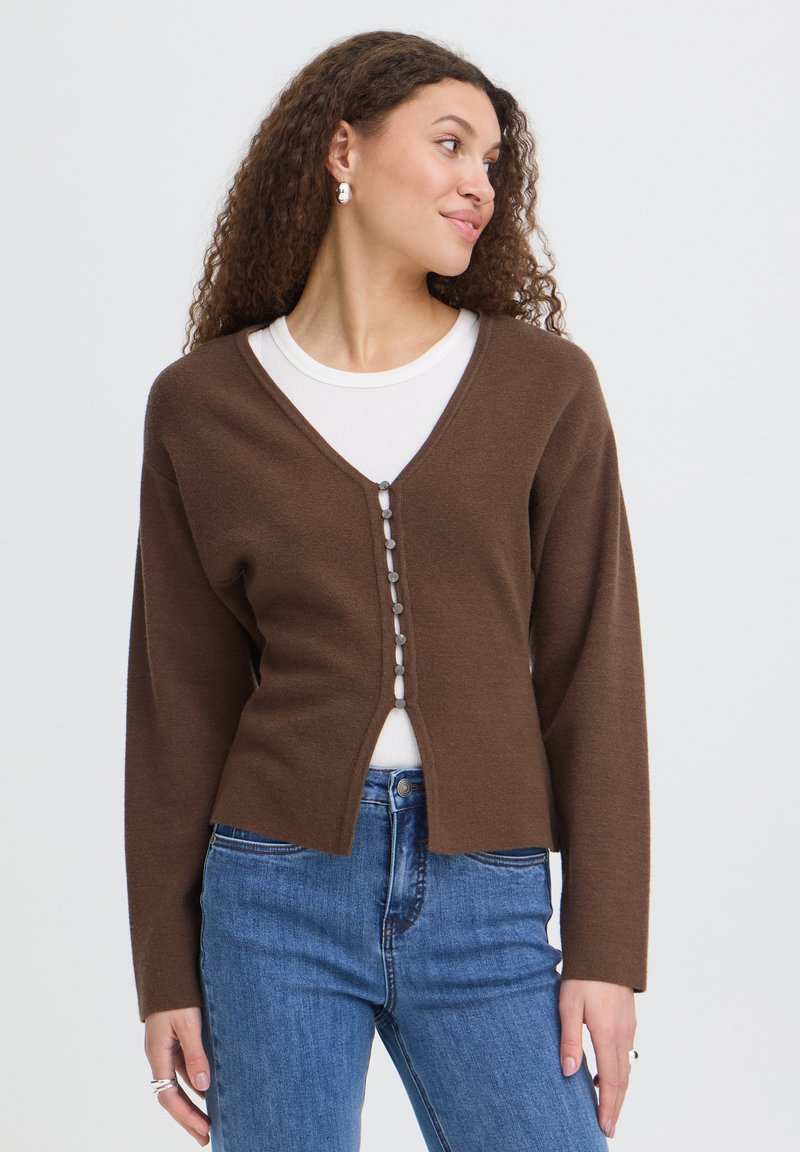 Femme aux cheveux bouclés portant un cardigan marron boutonné sur une chemise blanche et un jean bleu, regardant sur le côté devant un fond uni.