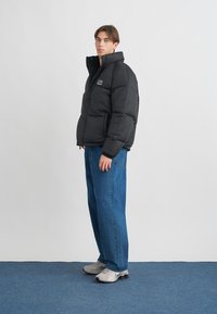 Μαύρο μπουφάν τύπου puffer με όρθιο γιακά, καπιτονέ σχέδιο, δύο τσέπες μπροστά, φορεμένο πάνω από μπλε τζιν παντελόνι φαρδιάς γραμμής και ανοιχτογρι αθλητικά παπούτσια.