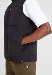 Black quilted vest with a high collar, front pockets, and a square pattern. Worn over a white short-sleeve shirt. Textured fabric.