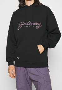 Černá mikina s kapucí, která má grafický textový design s nápisy "grimey" a "MUD CITY" v mnoha barvách vyšitými. Obsahuje přední kapsu a nastavitelnou kapuci.