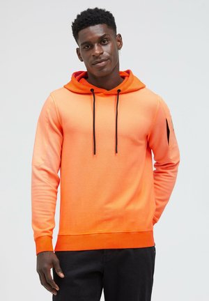 Junger Mann mit kurzen lockigen Haaren, der einen leuchtend orangefarbenen Hoodie mit schwarzen Kordeln und schwarze Hose trägt, vor einem einfarbigen hellen Hintergrund.