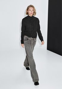 Cardigan grigio scuro con bottoni, jeans chiari svasati e calzature nere, con una texture liscia e un design minimalista su uno sfondo neutro.