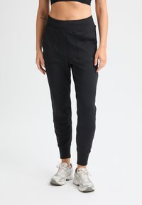 Pantalones deportivos negros con una cintura alta, tejido suave, perneras ajustadas y dos bolsillos frontales. Textura suave con puños elásticos.