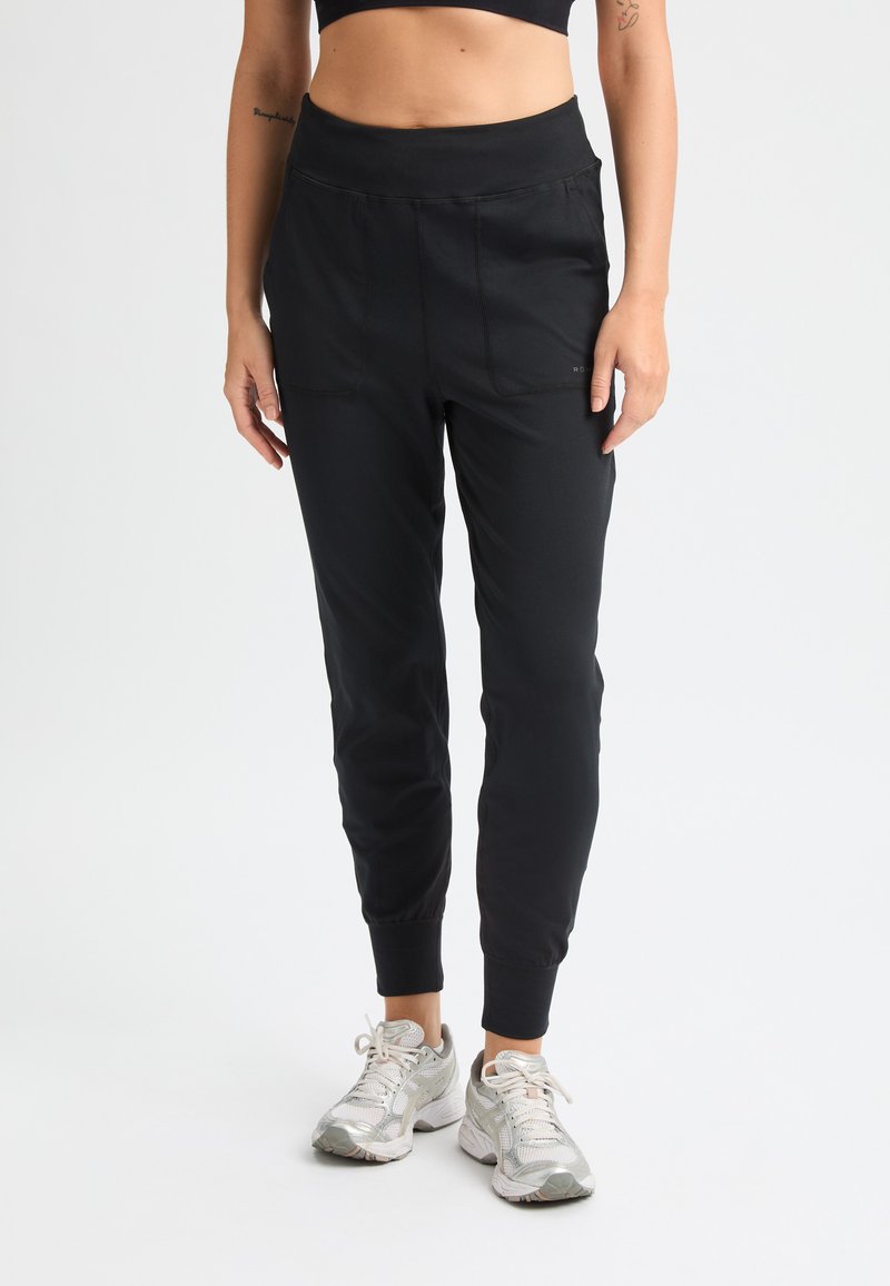 Pantalones deportivos negros con una cintura alta, tejido suave, perneras ajustadas y dos bolsillos frontales. Textura suave con puños elásticos.