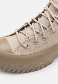 Zbliżenie na beżowy skórzany trampki z grubą gumową podeszwą i skrzyżowanymi sznurowadłami przewleczonymi przez metalowe przelotki.