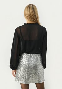 Blusa nera trasparente a maniche lunghe con una vestibilità svolazzante abbinata a una mini gonna argentata con paillettes, che riflette la luce con una superficie texturizzata.