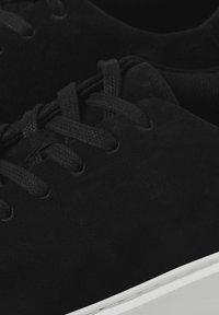 Baskets en daim noir avec des lacets assortis, dotées d'une semelle en caoutchouc blanc. La partie supérieure est lisse avec une texture subtile et des perforations.