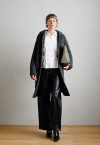 Cardigan grigio testurizzato, blusa bianca, pantaloni a gamba larga in pelle nera e una borsa verde. La modella si trova su un pavimento di legno di fronte a uno sfondo neutro.