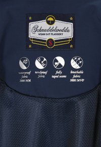 Tengerészkék vízálló dzseki, amelyen jól láthatóan hímzett márkacímke található, az ikonok pedig a vízálló, szélálló és lélegző anyagminőségeket jelzik.