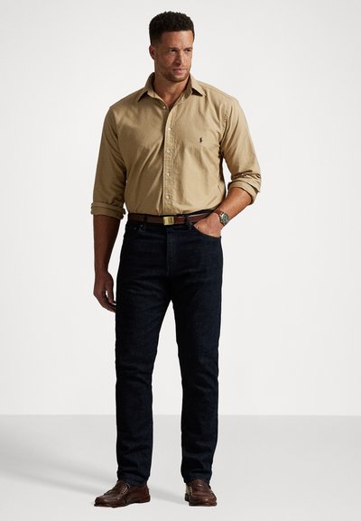 Beige gombos ing feltekert ujjakkal, sötétkék, testhezálló farmernadrág, barna bőr öv és barna mokaszin, az ingen logóval.