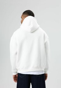 Witte hoodie met een losse pasvorm, zachte textuur, geribbelde boorden aan mouwen en zoom, gedragen over donkerblauwe spijkerbroek. Achteraanzicht.