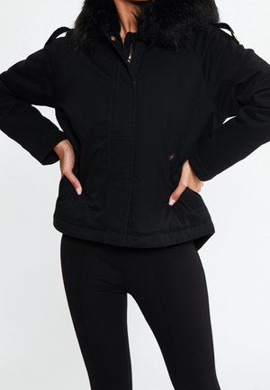 Femme portant une veste noire à col en fourrure, mains dans les poches, associée à des leggings noirs sur un fond blanc uni.