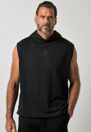 Gespierde man met baard, gekleed in een zwarte mouwloze hoodie met kap omlaag, staand tegen een effen lichte achtergrond, recht voor zich uitkijkend.