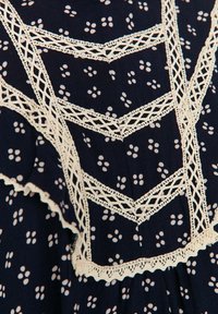 Tissu foncé avec de petits motifs floraux blancs à quatre points et une bordure de dentelle crème délicate en croisillons avec des motifs en chevrons et verticaux.