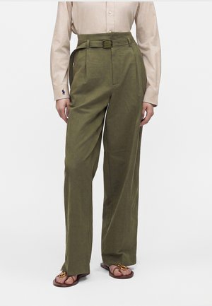 Mannequin portant un pantalon large taille haute vert olive avec ceinture assortie, chemise beige à manches longues et sandales marron avec détails dorés.