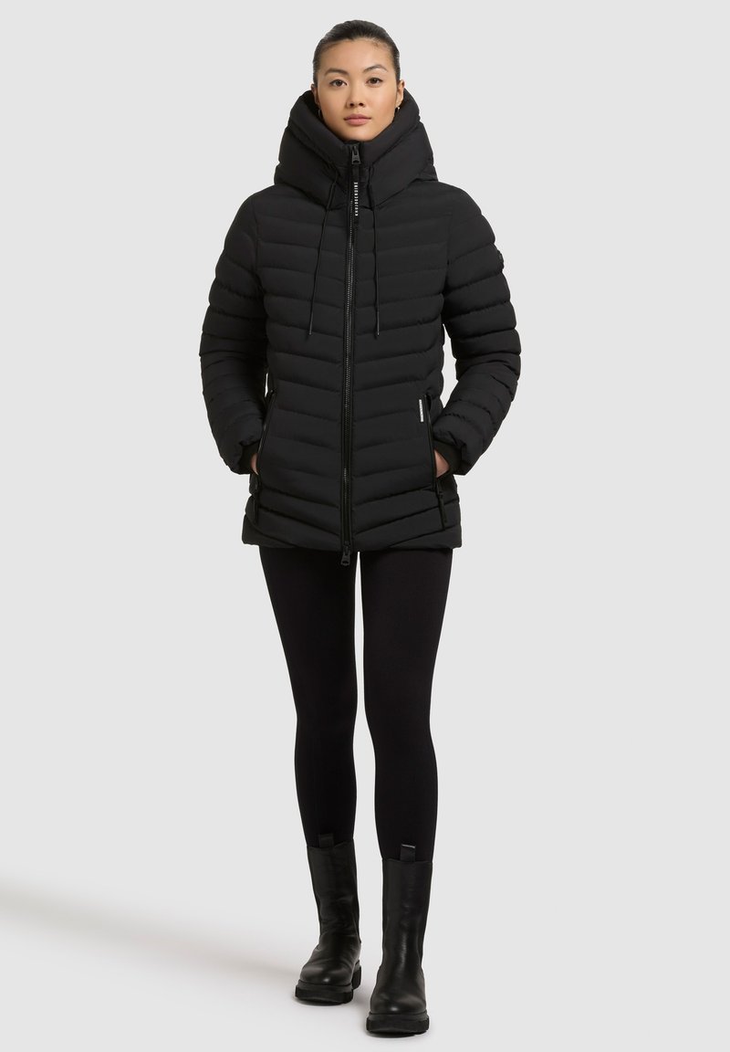 Giacca a vento nera con colletto alto e cappuccio regolabile, caratterizzata da un design a righe orizzontali e chiusura con zip. Abbinata a leggins neri e stivali.