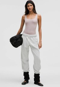 Body rose clair, pantalon de survêtement gris avec taille réglable, et jambières noires ; modèle tenant un sac noir, mettant en valeur des vêtements de sport décontractés.