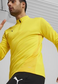 Top atletico giallo con colletto zip, realizzato in tessuto morbido. Presenta un logo nero e pannelli in rete ai lati per la traspirabilità.