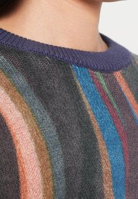 Gros plan sur une personne portant un pull en tricot à rayures verticales orange, vertes, bleues, roses et violettes, avec un col uni violet.