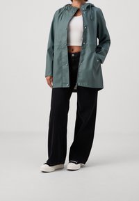 Waterbestendig turquoise regenjack met capuchon, drukknopen en zijzakken, gecombineerd met zwarte broek met wijde pijpen en witte sneakers.