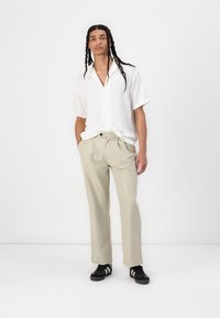 Camicia corta bianca a maniche corte con texture, pantaloni beige e sneakers nere con strisce bianche. Vestibilità casual, design semplice, colori solidi.
