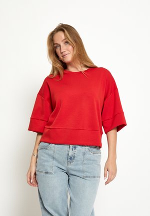 Vrouw met lang blond haar, gekleed in een losse rode top met korte mouwen en lichtblauwe jeans met hoge taille, staand tegen een witte achtergrond.