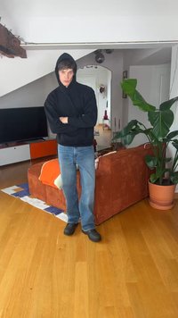 Νέος άνδρας με μαύρο φούτερ με κουκούλα και τζιν στέκεται με σταυρωμένα χέρια σε ένα σαλόνι με πορτοκαλί καναπέ, μεγάλο φυτό, τηλεόραση και ξύλινο πάτωμα.