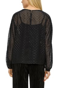 Schwarze transparente Bluse mit gepunkteten Verzierungen, langen Ärmeln und einem Schlüssellochverschluss auf der Rückseite. Kombiniert mit plissierten schwarzen Hosen.