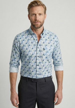 Man met kort blond haar en baard, dragend een lichtblauw bloemmotief overhemd met opgerolde mouwen en een donkere broek, staand tegen een effen achtergrond.