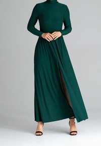 Maxikleid mit langen Ärmeln und hohem Kragen aus tiefgrünem Stoff, mit seitlichem Schlitz, glatter Textur und kombiniert mit schwarzen Riemchen-Heels.