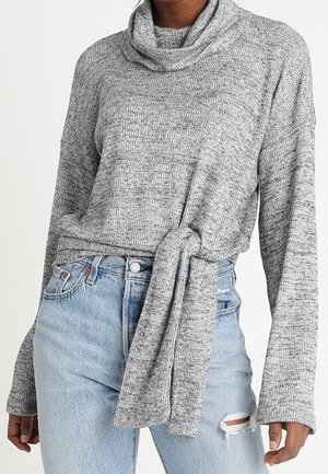 Maglione grigio a collo alto con texture e maniche lunghe, con un dettaglio annodato in vita, abbinato a jeans strappati di un blu chiaro.