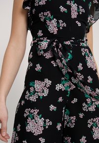 Černé květinové šaty s růžovými a bílými květy a zelenými listy, s volánovými rukávy a detailem vázáním v pase. Měkká tkanina s lehkým a splývavým textura.