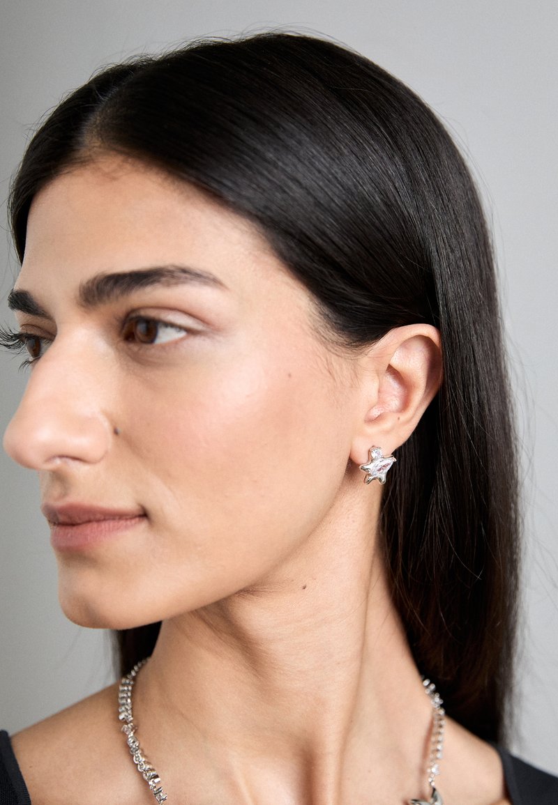 Ασημένιο σκουλαρίκι σε σχήμα αστεριού με γυαλιστερή φινίρισμα. Κοντινή άποψη που τονίζει τις λεπτομέρειες του σχεδίου και την υφή, σε αντίθεση με ουδέτερο φόντο.