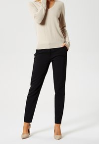 Femme portant un pull beige à encolure en V, un pantalon noir ajusté et des escarpins beiges, debout avec une main dans la poche.